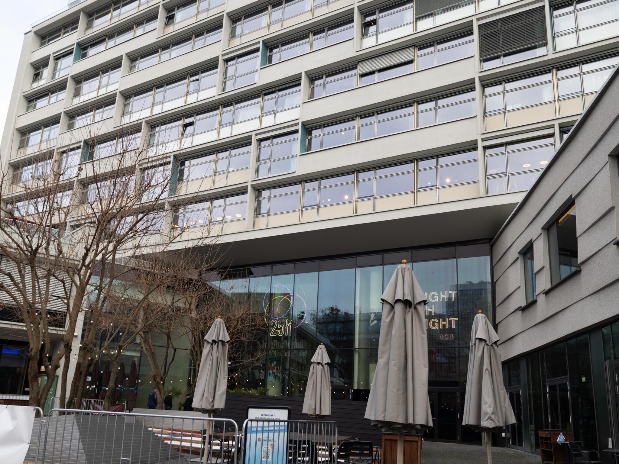 Fassade des 25hours Hotels mit klarer, moderner Linienführung und zahlreichen bodentiefen Fenstern. Im unteren Bereich befindet sich der gläserne Eingangsbereich mit dem markanten 25hours-Logo, daneben eine Außenterrasse mit geschlossenen Sonnenschirmen. Kahle Bäume im Vordergrund vermitteln eine winterliche Stadtszenerie, während die Architektur des Hotels urban und zeitgemäß wirkt.